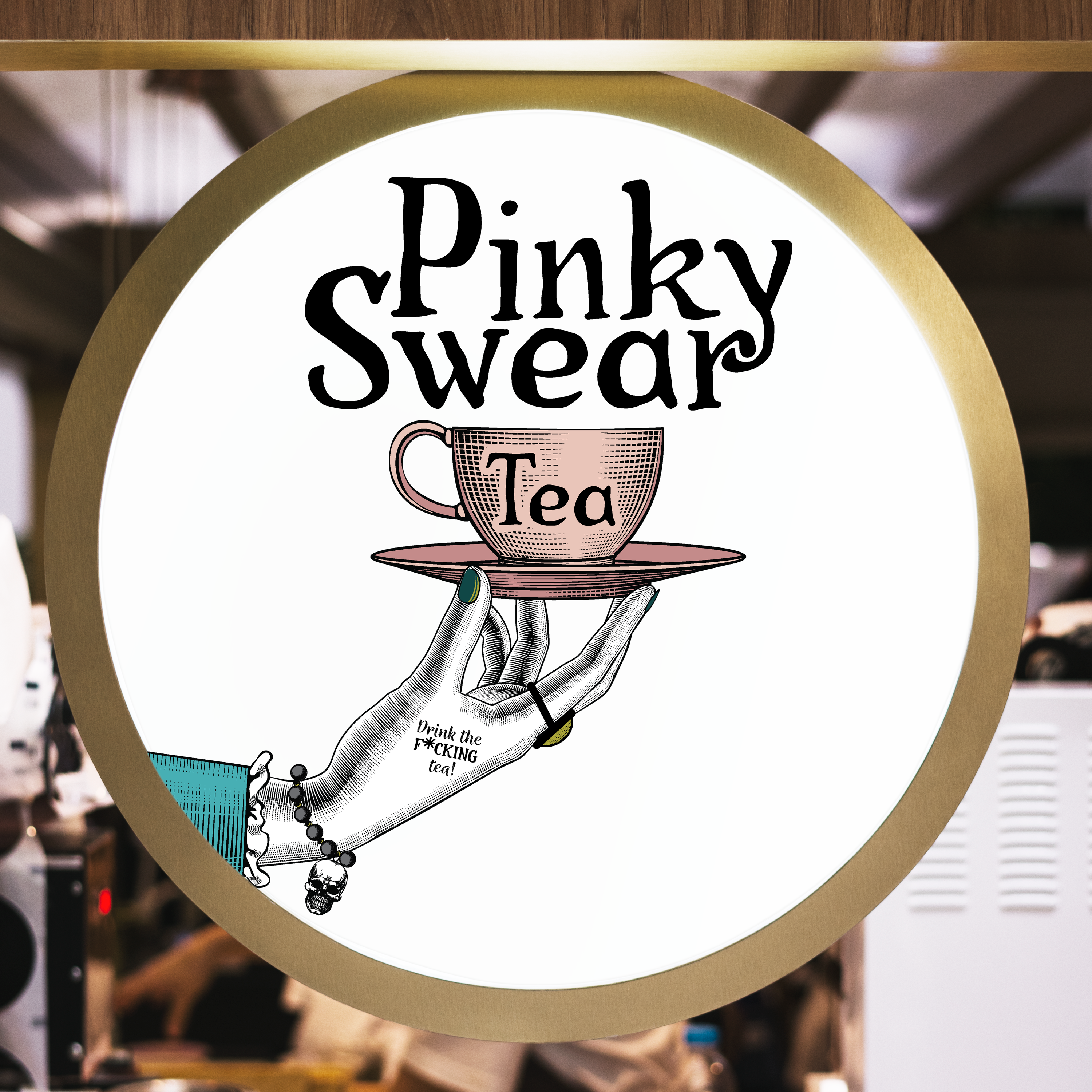 A circular sign featuring a hand holding a teacup on a saucer with the text 'Pinky Swear Tea' and the phrase 'Drink the F*CKING tea!' in a playful font.